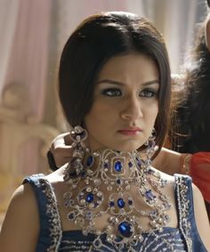 a woman in a blue dress is getting her hair done by another woman wearing an elaborate necklace and earrings