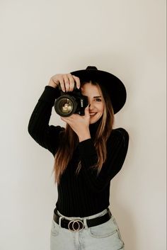 a woman wearing a black hat and holding a camera up to her face with both hands