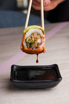 a person holding chopsticks above a sushi roll on a black plate with sauce drizzled over it