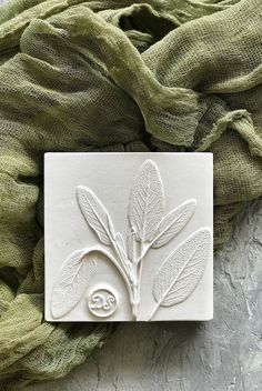 a white tile with leaves on it sitting next to a green cloth and a piece of fabric