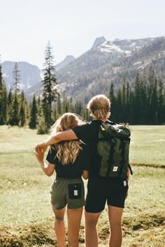 two people with backpacks are standing in the grass looking at mountains and pine trees