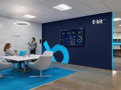 two women are talking in an office with blue and white accents on the walls, while one woman is standing at a table