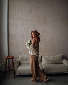 a woman standing in front of a couch holding a bouquet of flowers and looking at the wall