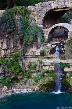 a waterfall with a tunnel in the middle surrounded by greenery and stone walls next to a body of water