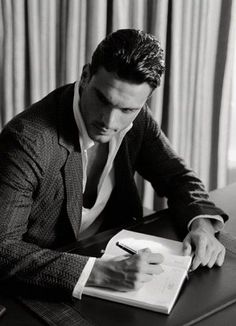 a man sitting at a desk writing on a book with a pen and paper in front of him