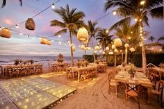 an outdoor dining area on the beach with lights strung from palm trees and hanging lanterns