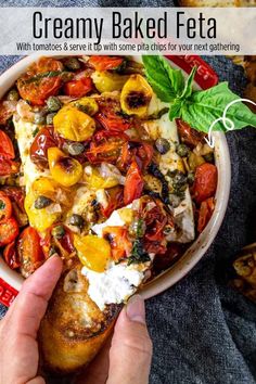 a person holding a piece of bread with tomatoes and olives on it in front of a bowl of baked feta