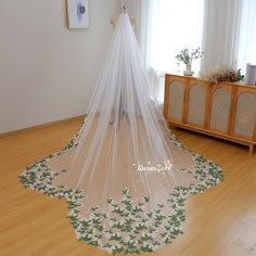 a white wedding veil with green leaves on the bottom is hanging in front of a window