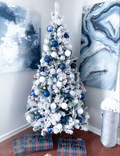 a white christmas tree with blue and silver ornaments in front of two paintings on the wall