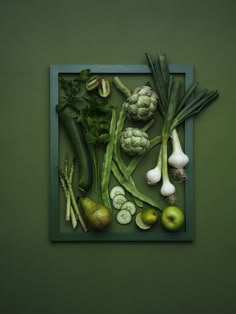 an arrangement of vegetables displayed in a square frame on a green wall, including broccoli, onions, cucumbers and pears