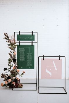 two black metal stands with flowers on them against a white brick wall, one holding a menu and the other displaying an event plan