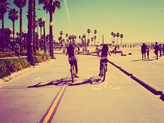 black and white photograph of two people riding bikes on the street with palm trees in the background