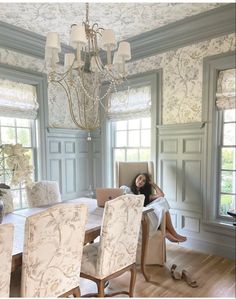 a woman sitting at a dining room table in front of a chandelier and windows