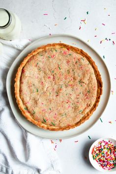 a sprinkled pie on a white plate next to a glass of milk and some sprinkles