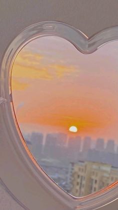 a heart shaped window with the sun setting in the distance and buildings seen through it