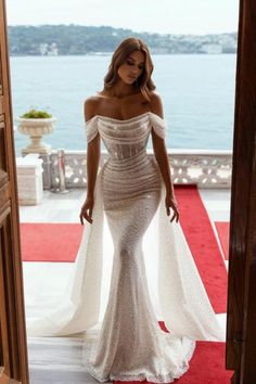 a woman in a white dress standing on a red carpet near the water with her arms behind her back