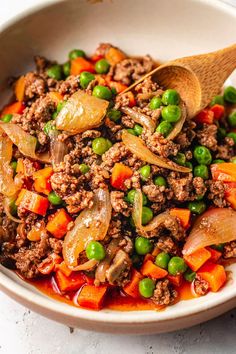 a white bowl filled with meat, peas and carrots next to a wooden spoon