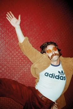 a man with his hands in the air while sitting on a red wall wearing adidas t - shirt