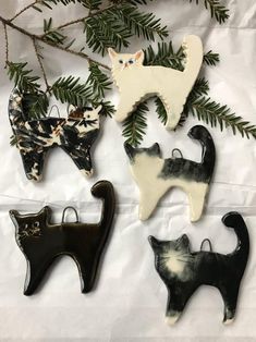 four ceramic cats hanging from a tree branch on a white sheet with pine branches in the background