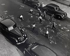 black and white photograph of people playing with chalk on the sidewalk in front of parked cars