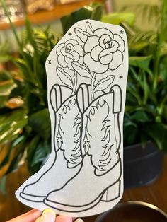 a person holding up a paper cutout with boots and flowers on it in front of a potted plant