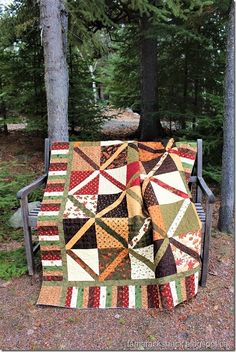 a patchwork quilt sitting on top of a bench in front of some pine trees