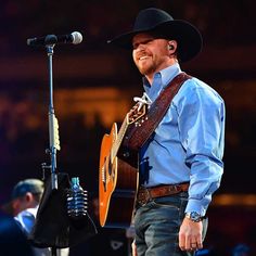 a man wearing a cowboy hat and holding a guitar in front of a microphone on stage