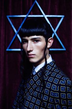 a man with long black hair wearing a patterned jacket and standing in front of a star of david