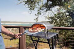 a man holding a pizza on top of an outdoor grill with the ocean in the background