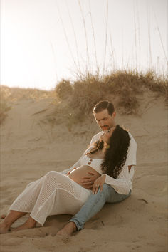 a man and woman are sitting in the sand together while holding each other's belly