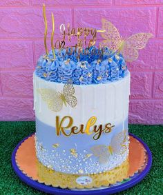 a birthday cake decorated with blue flowers and gold butterflies is on a plate in front of a pink brick wall