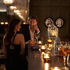 a man and woman sitting at a bar with candles in front of them, talking to each other