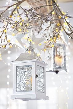 two white lanterns hanging from a tree with lights in the background and snow flakes on the branches