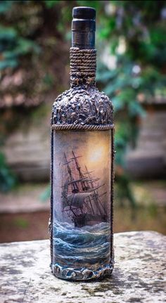 a bottle with a painting on it sitting on a rock next to some water and trees