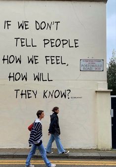 two people walking down the street in front of a white wall with writing on it