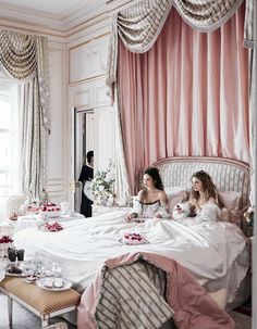 two women sitting on a bed with pink drapes over them and one holding a cup of coffee