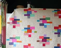 a white quilt with multicolored squares is hanging on the wall next to a lamp