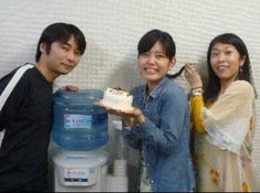 three people standing next to each other with a cake in front of them and a water cooler behind them