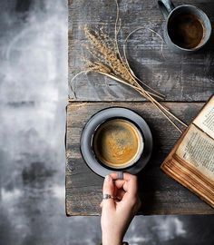 a person holding a cup of coffee on top of a table next to an open book