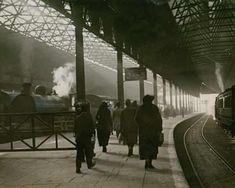 an old photo of people walking on the train tracks near a train station with steam pouring out of it