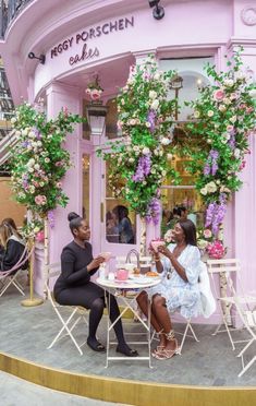 two women sitting at a table in front of a pink building with flowers on it