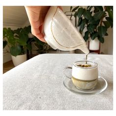 someone pouring something into a small cup on top of a white cloth covered table with potted plants in the background