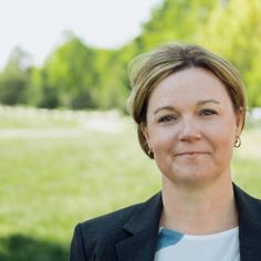 a woman standing in front of a green field with trees and grass behind her, wearing a black blazer