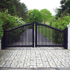 an iron gate and brick walkway leading to a driveway surrounded by greenery on either side