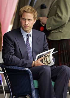 a man in a kilt sitting on a chair reading a magazine with other people standing behind him