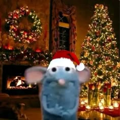 a blue stuffed animal wearing a santa hat in front of a christmas tree and fireplace