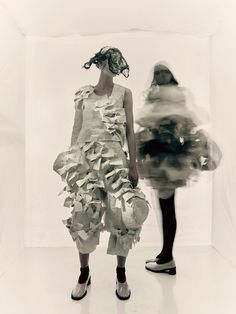 two women standing next to each other in front of a white wall with paper on it