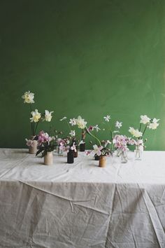 several vases with flowers on a table in front of a green wall and white cloth