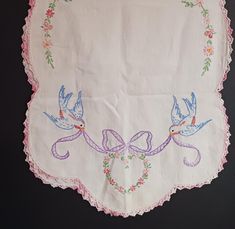 an embroidered tablecloth with two birds and ribbons on the border, hanging from a black wall