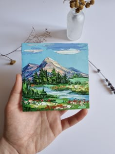 a hand holding up a small piece of art with mountains and trees in the background